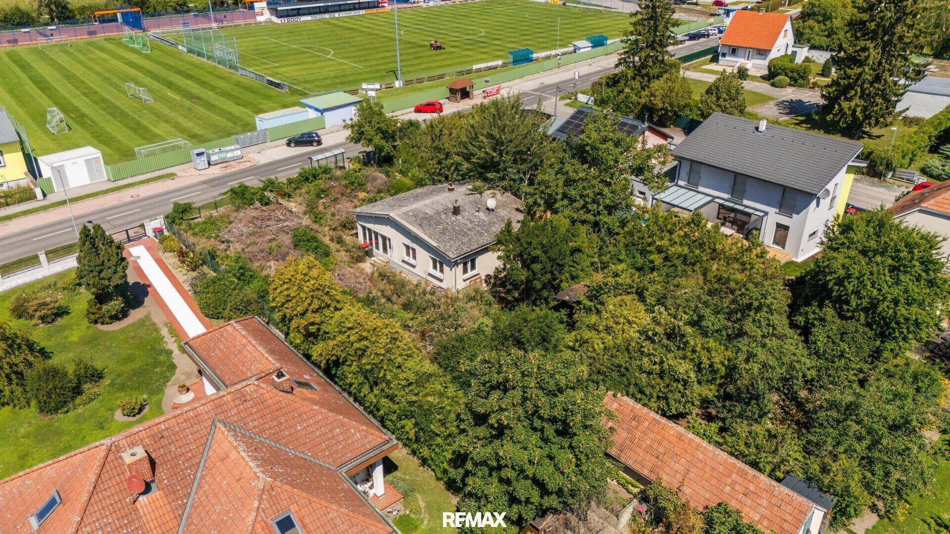 Luftaufnahme des Hauses in einer grünen Umgebung mit angrenzendem Sportplatz und Nachbarhäusern.