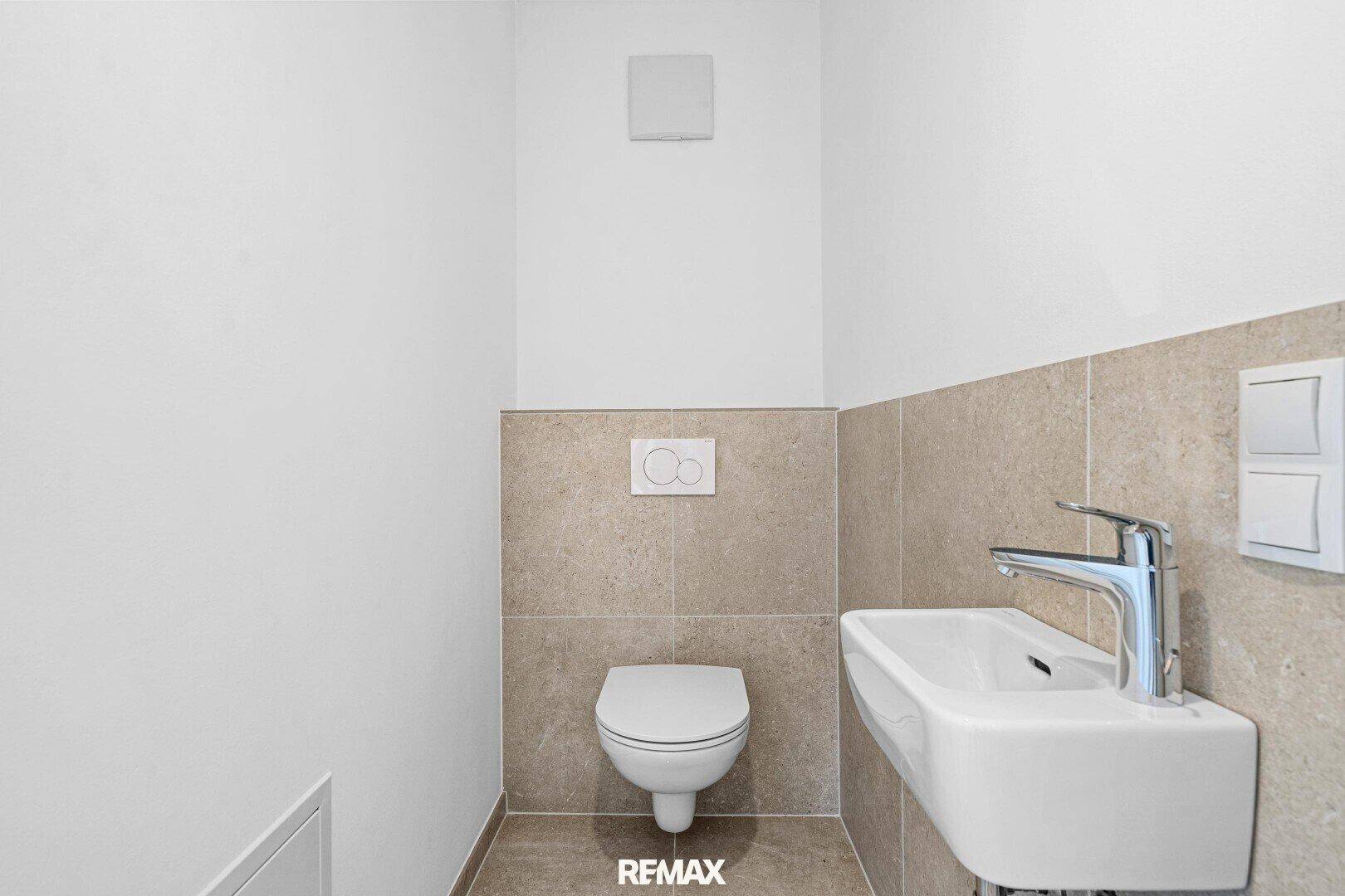 Gäste-WC mit Wand-WC, kleinem Waschbecken und beigefarbenen Fliesen an der Wand.