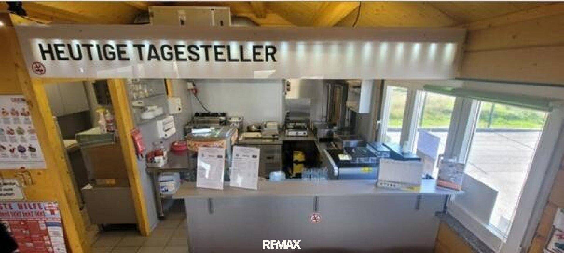 Innenansicht eines Imbisses mit Theke, Küchengeräten und einer beleuchteten Anzeigetafel.