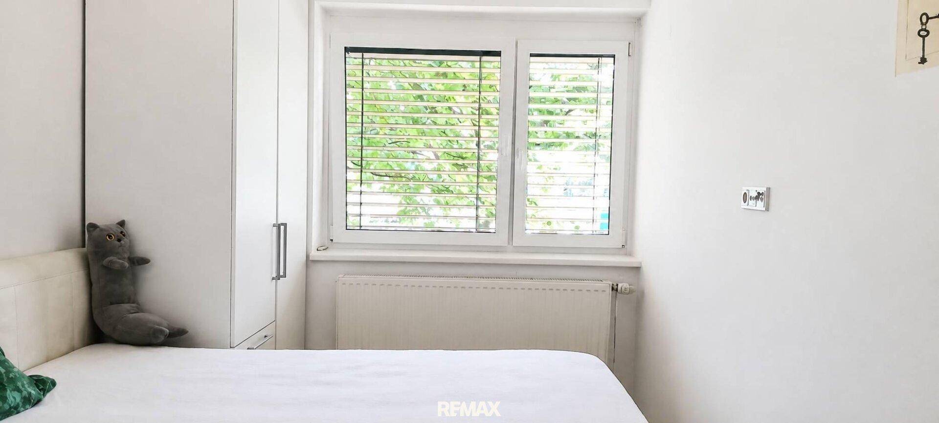 Helles Schlafzimmer mit weißem Bett, Kleiderschrank und Fenster mit Jalousien.