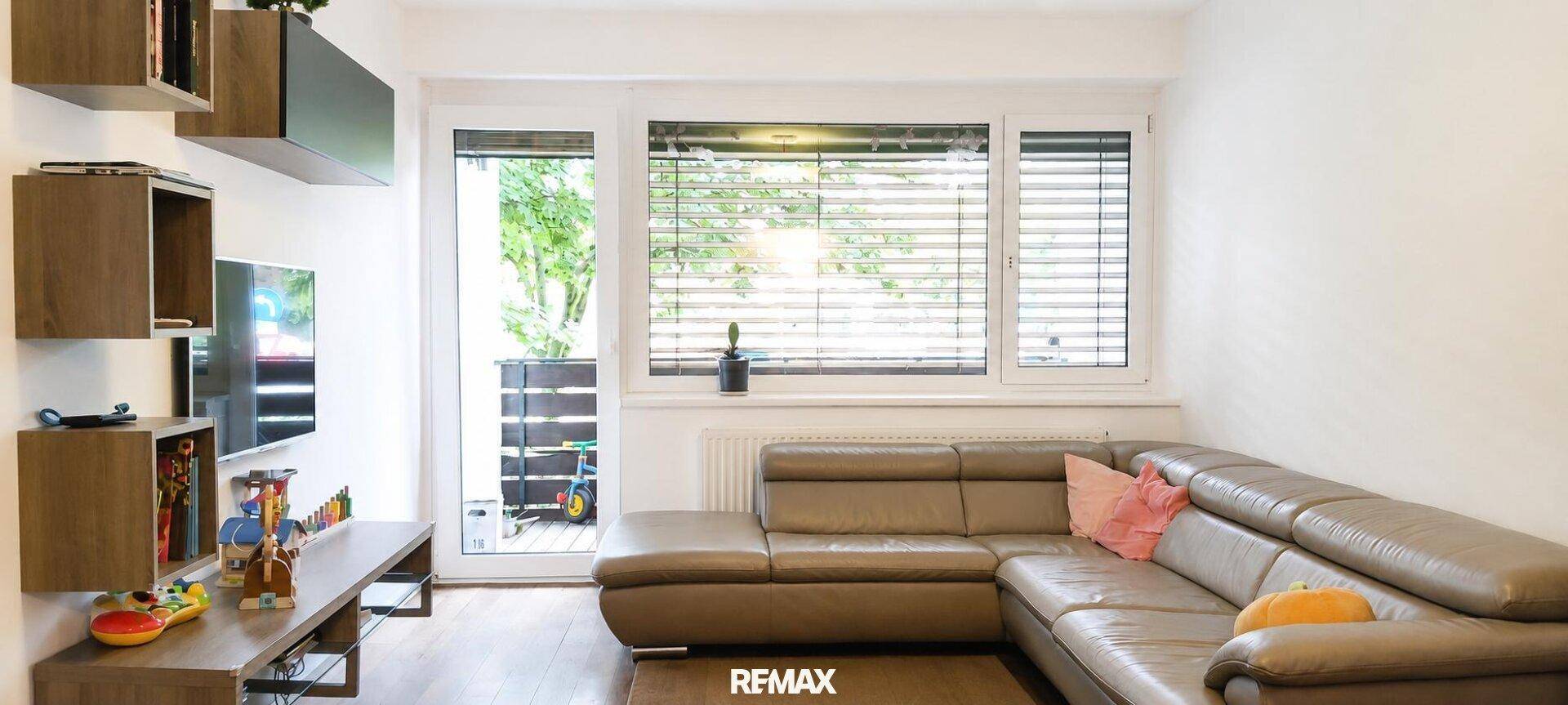 Gemütliches Wohnzimmer mit braunem Ecksofa, Holzregalen und Zugang zu einem Außenbereich.