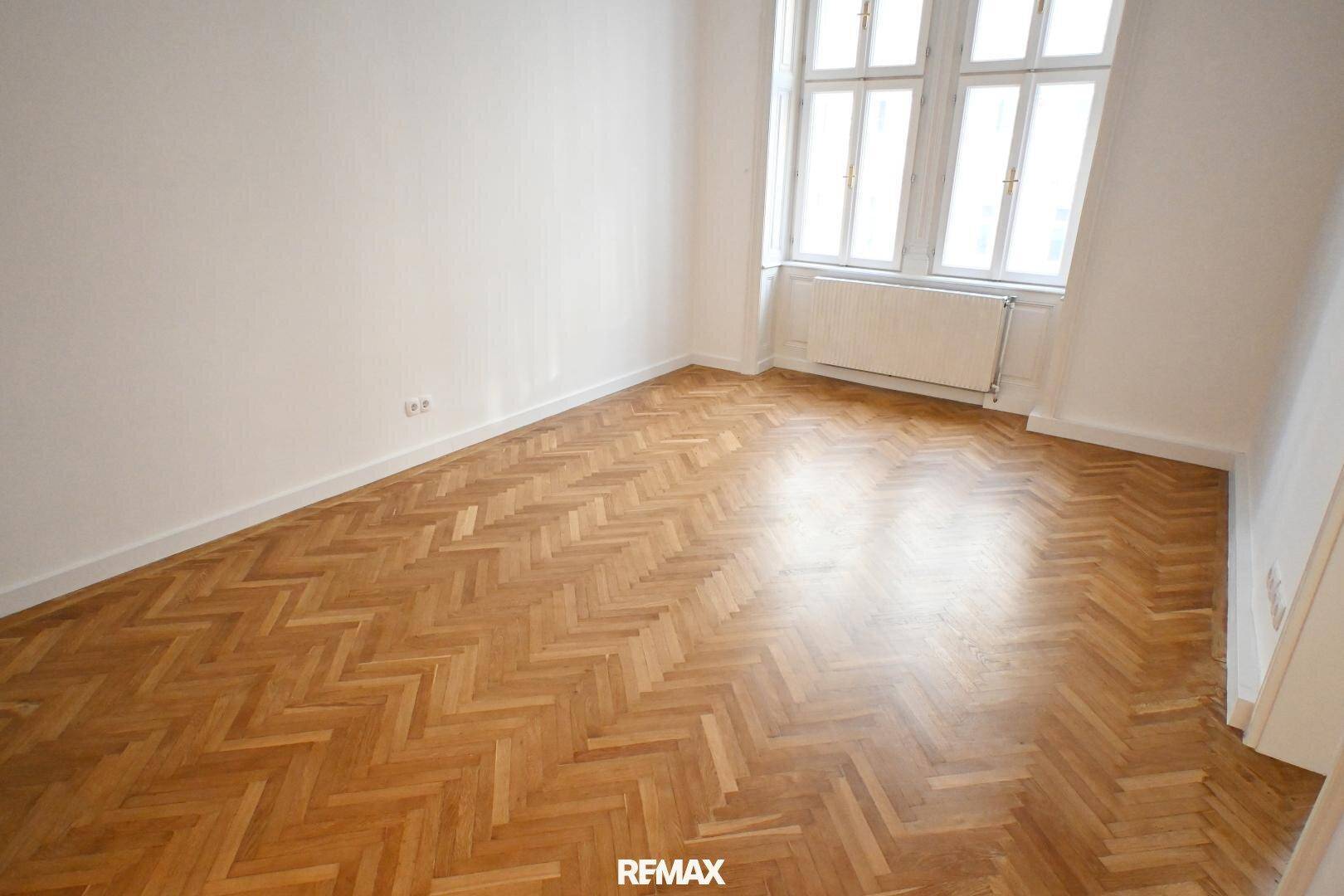Leeres Zimmer mit glänzendem Fischgrätparkett und zwei hohen Fenstern.