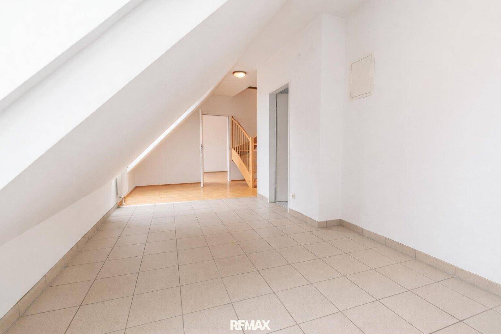 Leerer Raum im Dachgeschoss mit Fliesenboden, Dachschräge und Zugang zur Treppe.