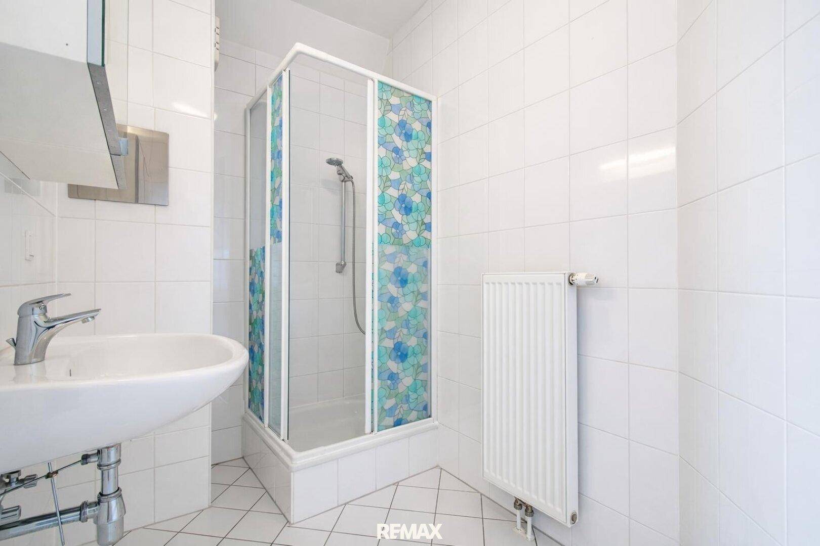 Helles Badezimmer mit Duschkabine, weißen Fliesen und Heizkörper als Handtuchhalter.