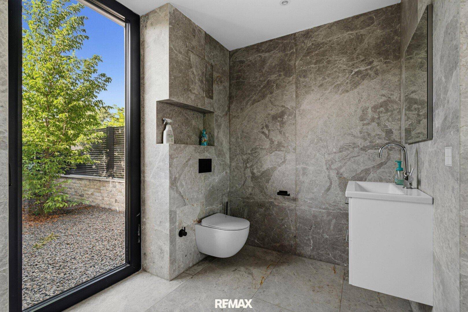 Modernes WC mit Marmorfliesen, wandmontierter Toilette und Fenster zum Garten.