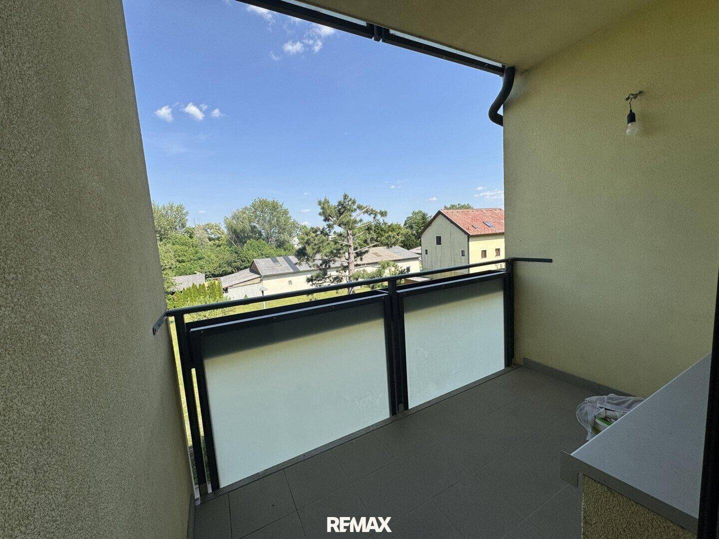 Balkon mit Fliesenboden und Blick auf umliegende Häuser und grüne Bäume unter blauem Himmel.