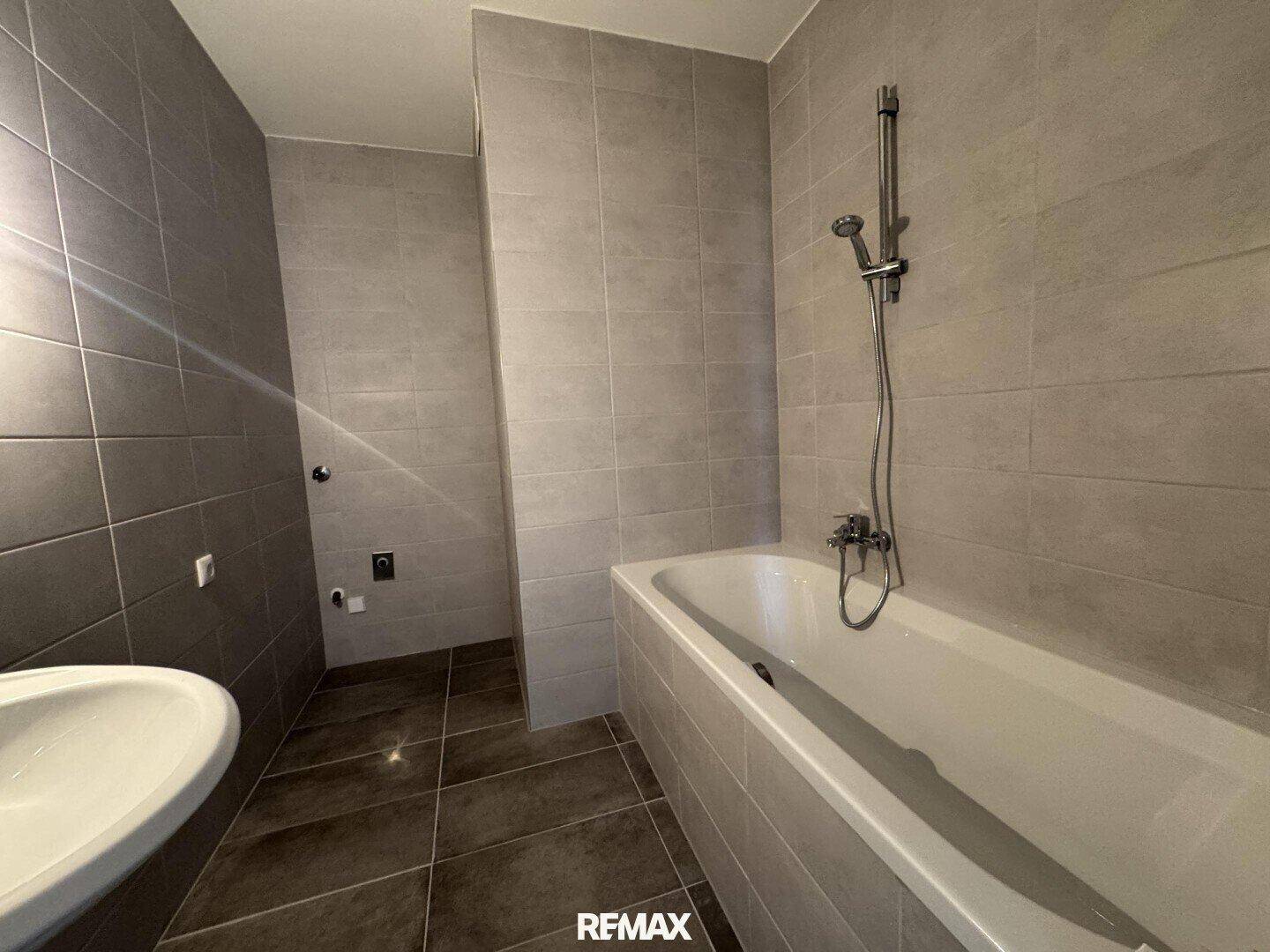 Modernes Badezimmer mit grauen Fliesen, einer Badewanne mit Duschfunktion und einem Waschbecken.