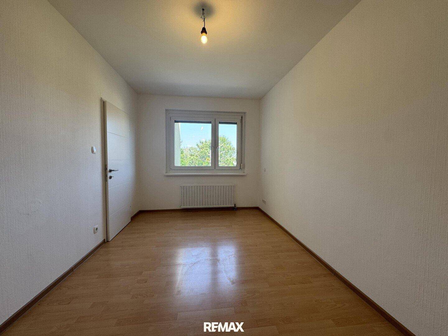 Leeres Schlafzimmer mit Holzboden, einem Fenster und einer weißen Tür, gut beleuchtet.