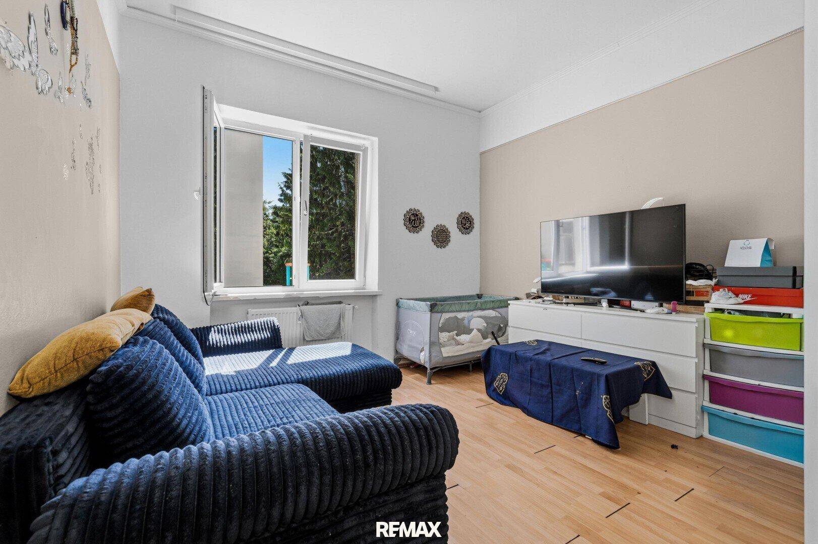 Wohnzimmer mit blauem Sofa, Fernseher, Kommode und Babybett.