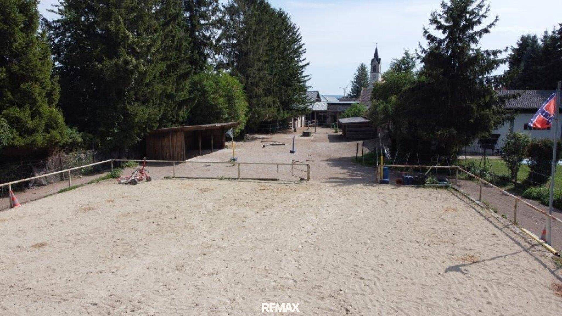 Weitläufiger, eingezäunter Sandplatz, der als Reitplatz genutzt werden könnte, mit ländlicher Umgebung.