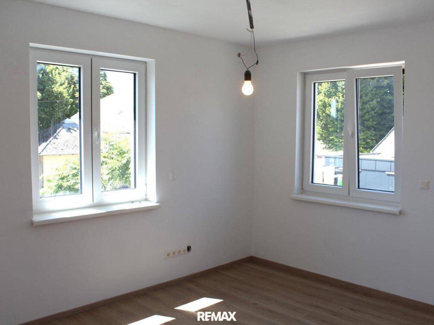 Leeres Zimmer mit zwei Fenstern, die einen grünen Ausblick bieten, und hellem Laminatboden.