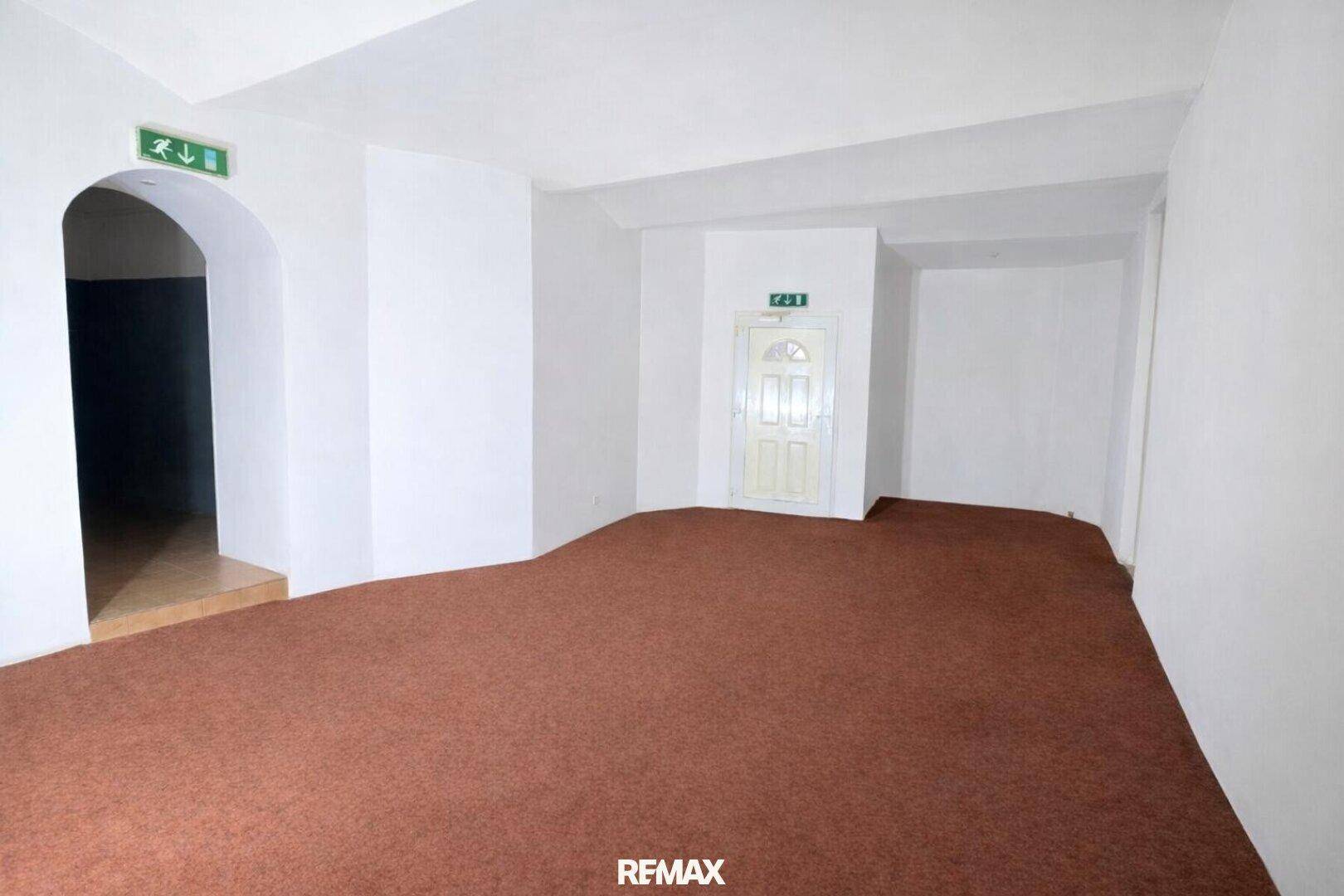 Großer, leerer Raum mit braunem Teppichboden und einem Durchgang zu einem weiteren Bereich.