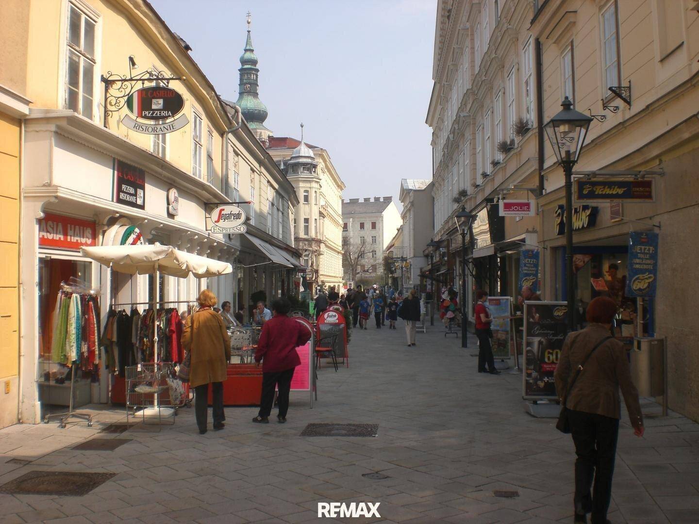 Belebtes Stadtbild mit Geschäften, Passanten und einem Kirchturm im Hintergrund, typisch für eine Einkaufsstraße.
