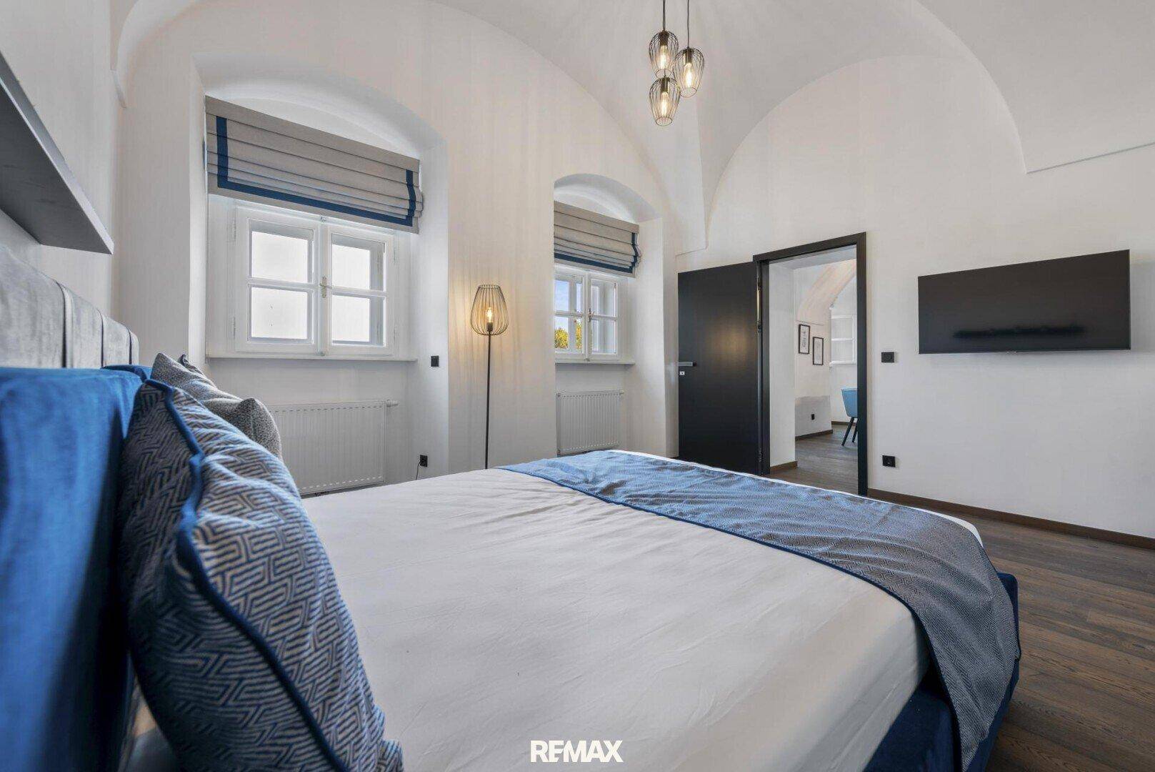 Komfortables Schlafzimmer mit Doppelbett, Fenstern und Zugang zu einem weiteren Raum.