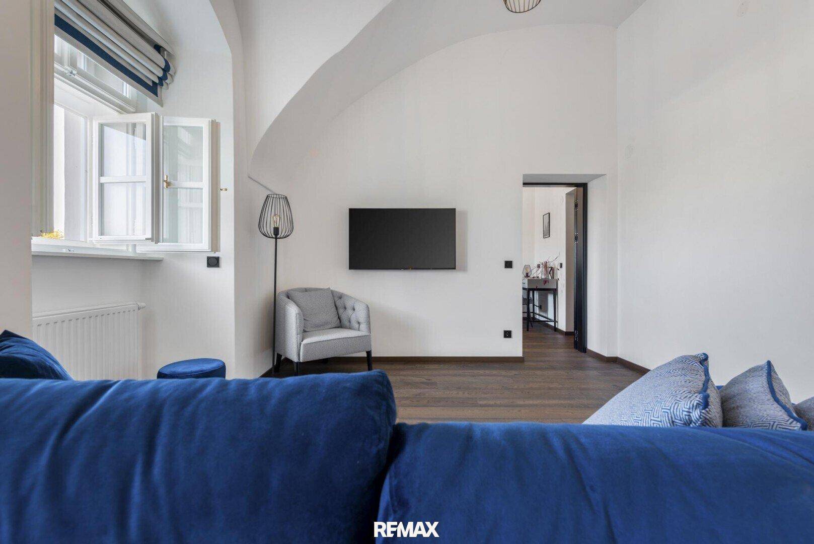 Gemütliches Wohnzimmer mit blauer Couch, Wandfernseher und natürlichem Lichteinfall durch das Fenster.