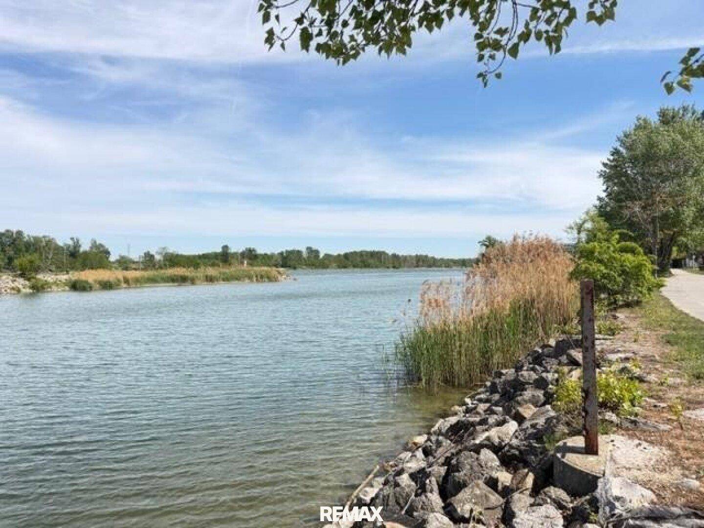 Ruhiges Gewässer mit Schilf und Felsen am Ufer unter blauem Himmel.