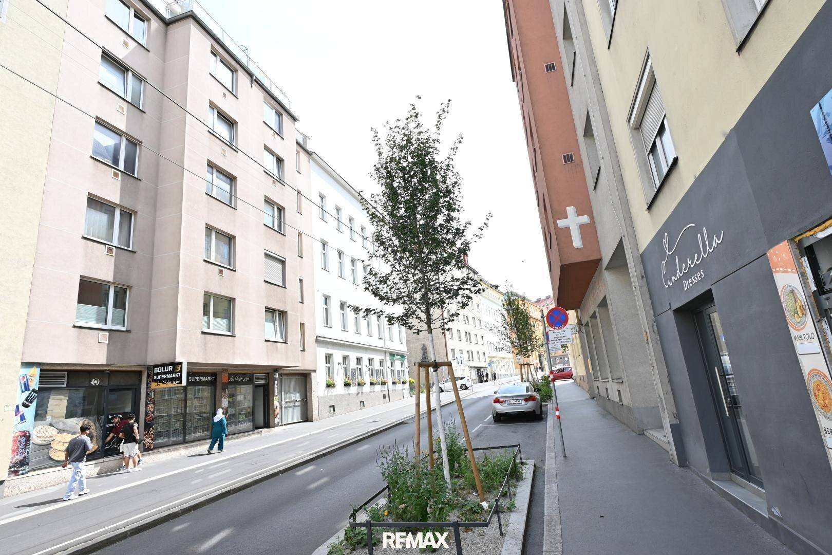 Straßenansicht mit Gebäuden, Bäumen und parkenden Autos in einer städtischen Umgebung.