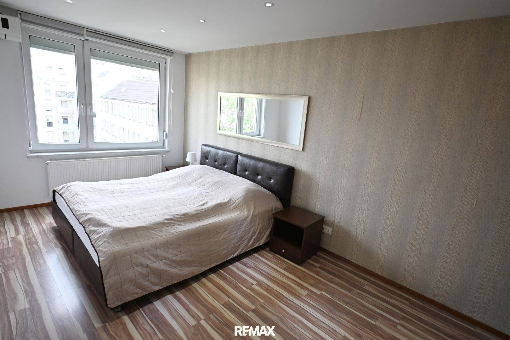 Schlafzimmer mit Doppelbett, Nachttisch und gemusterter Tapete, gut beleuchtet durch ein Fenster.