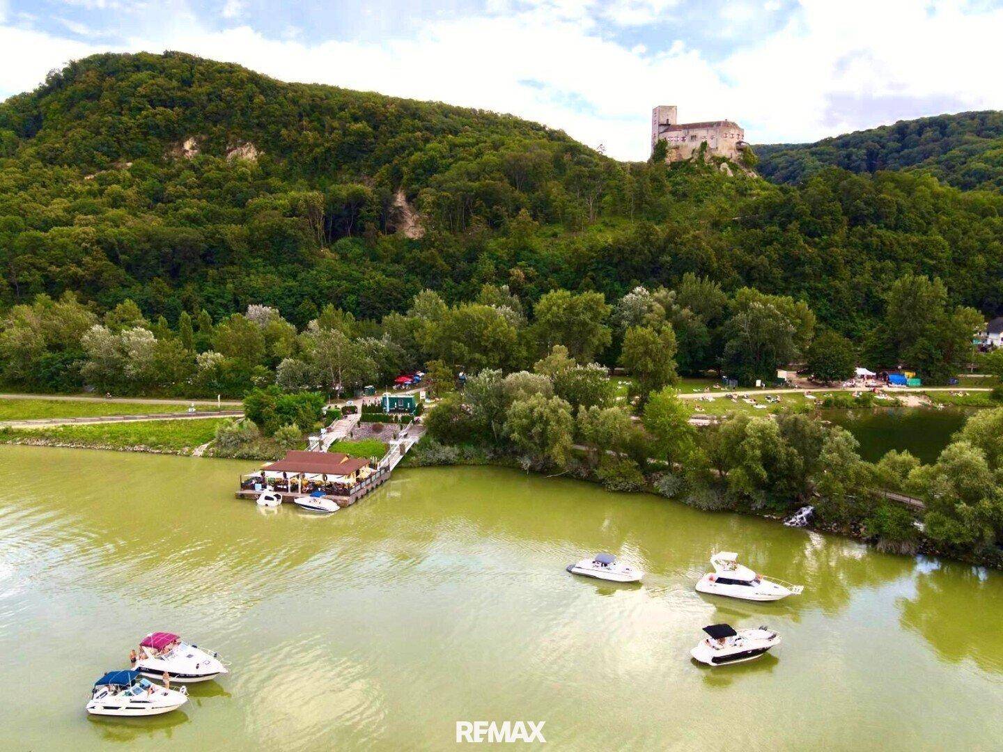 Weitläufige Luftaufnahme des Sees mit dem schwimmenden Gebäude, Booten und einer Burgruine auf einem bewaldeten Hügel.