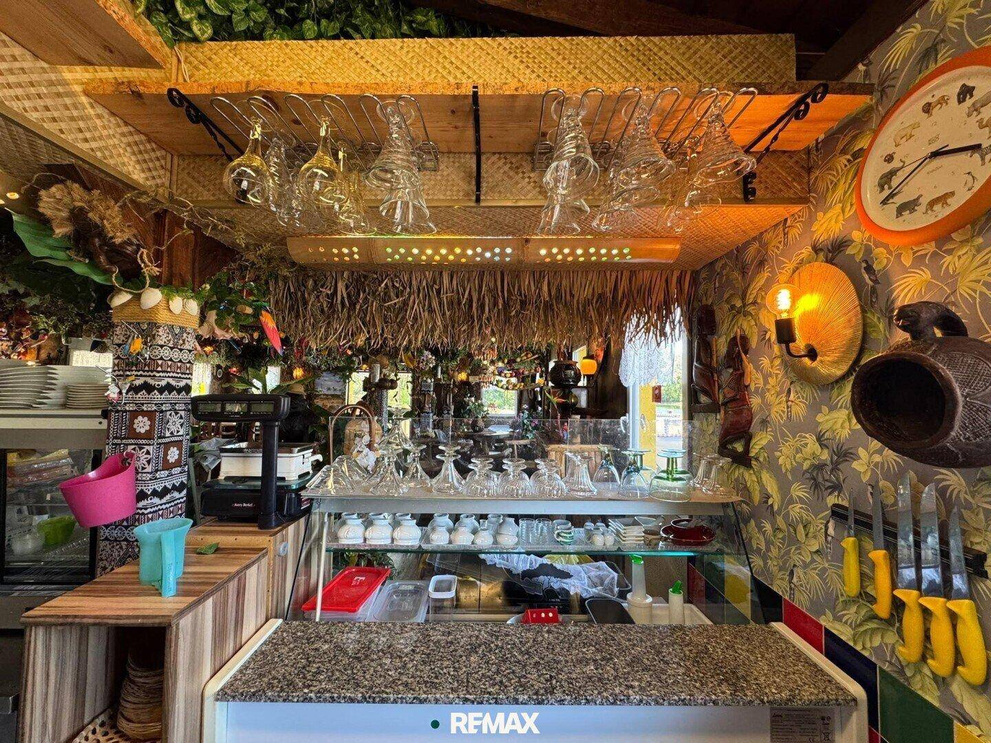 Thekenbereich mit Glasvitrine, Kaffeemaschine und hängenden Weingläsern unter einem Strohdach.