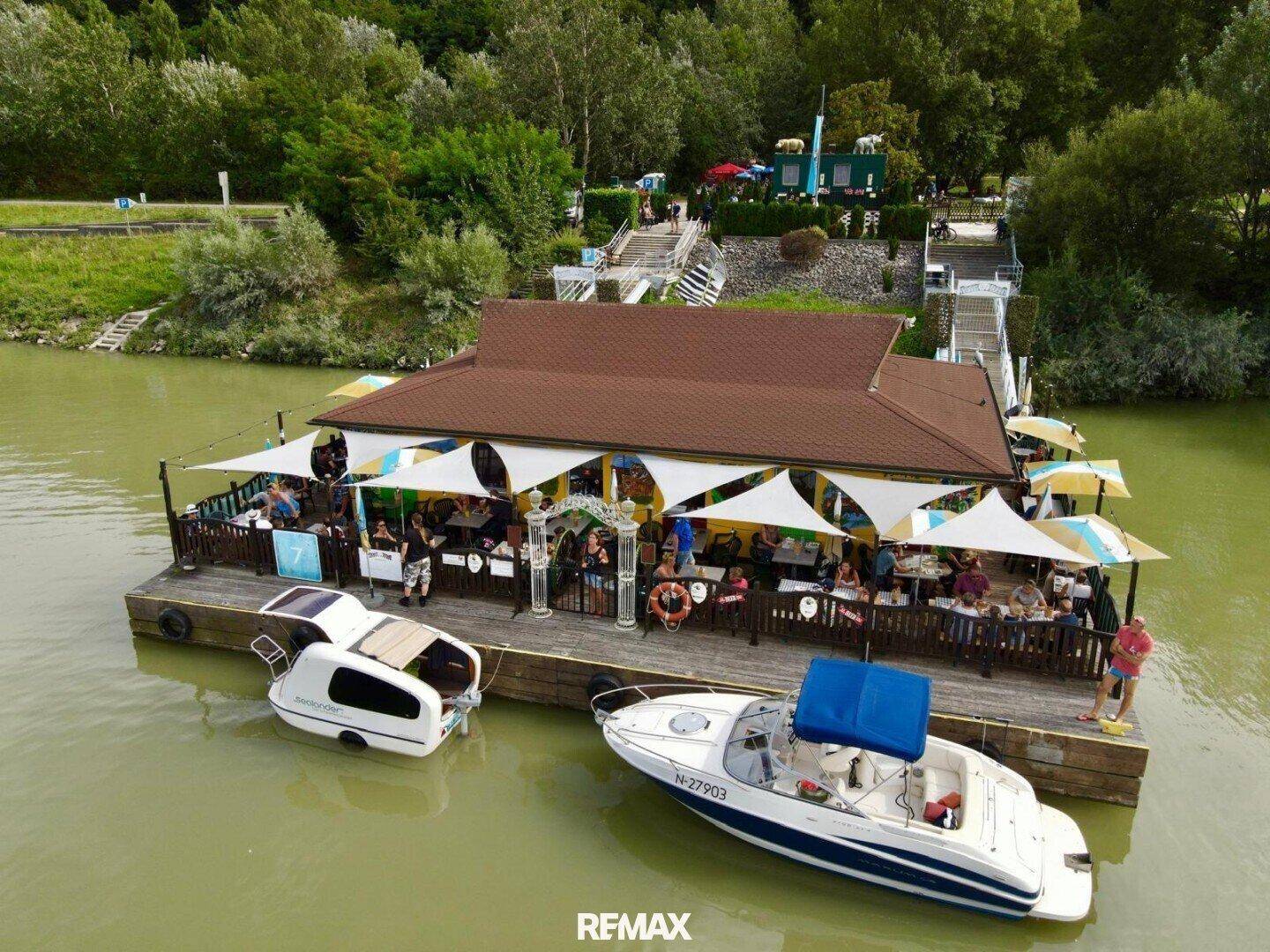 Eine Luftaufnahme des schwimmenden Restaurants mit Gästen auf der Terrasse und Booten am Anleger.