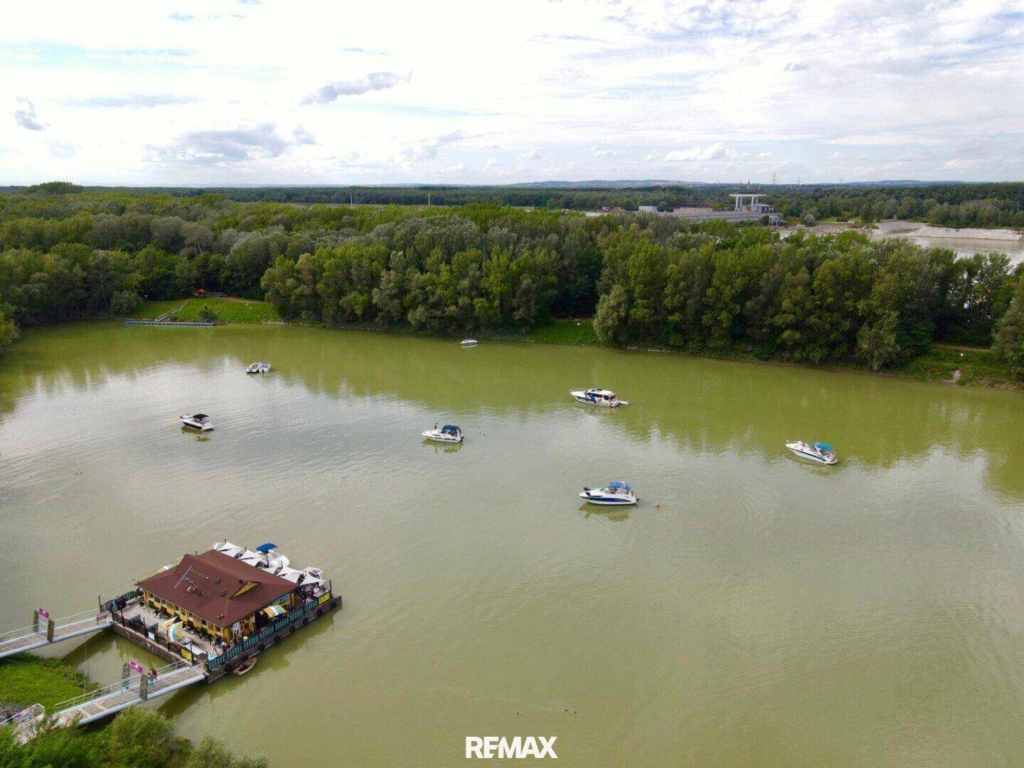 Weitläufige Luftaufnahme des Sees mit dem schwimmenden Gebäude und zahlreichen Booten, umgeben von Wald.