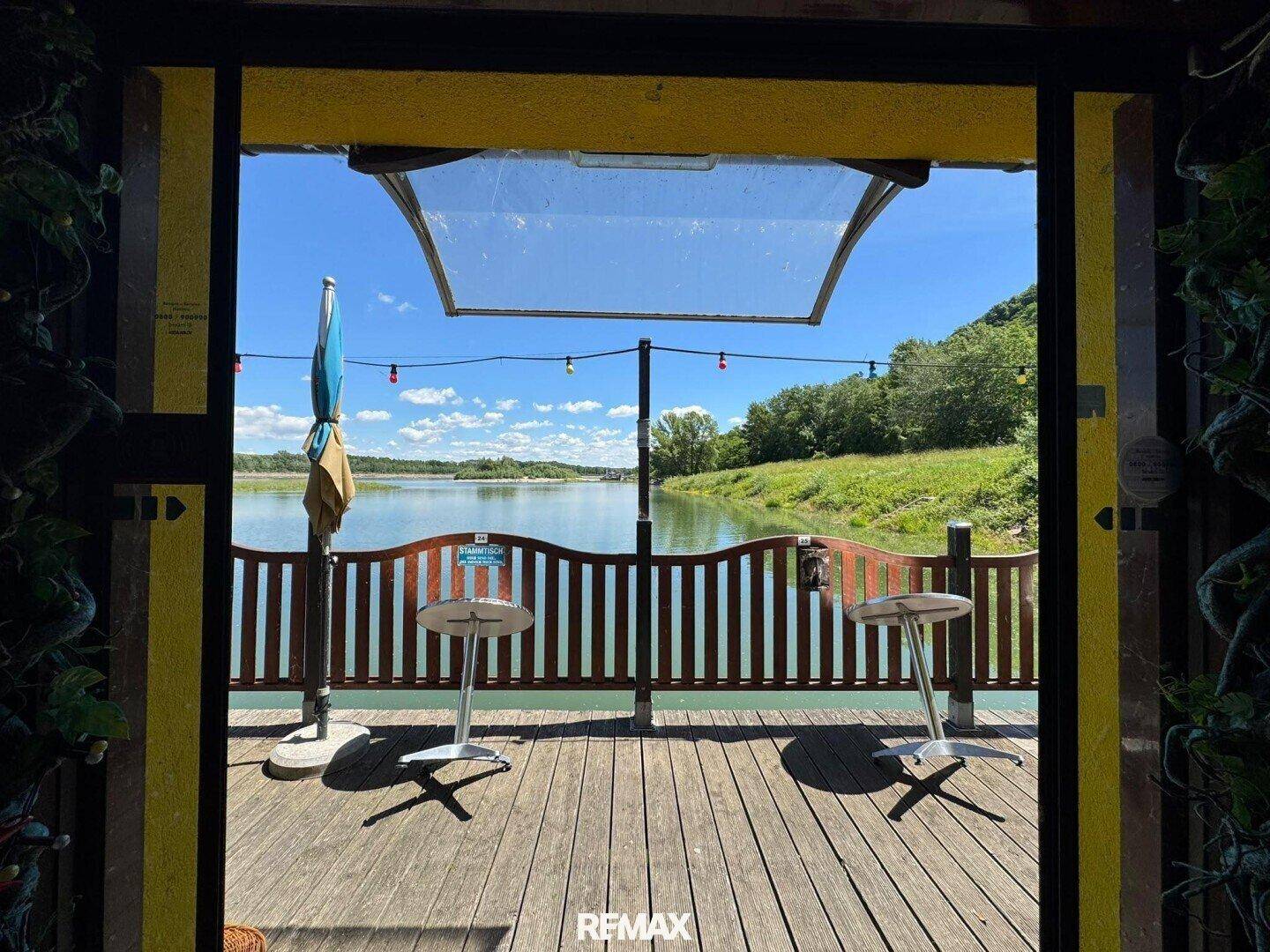 Blick von der Holzterrasse auf das ruhige Wasser und die grüne Uferlandschaft unter blauem Himmel.