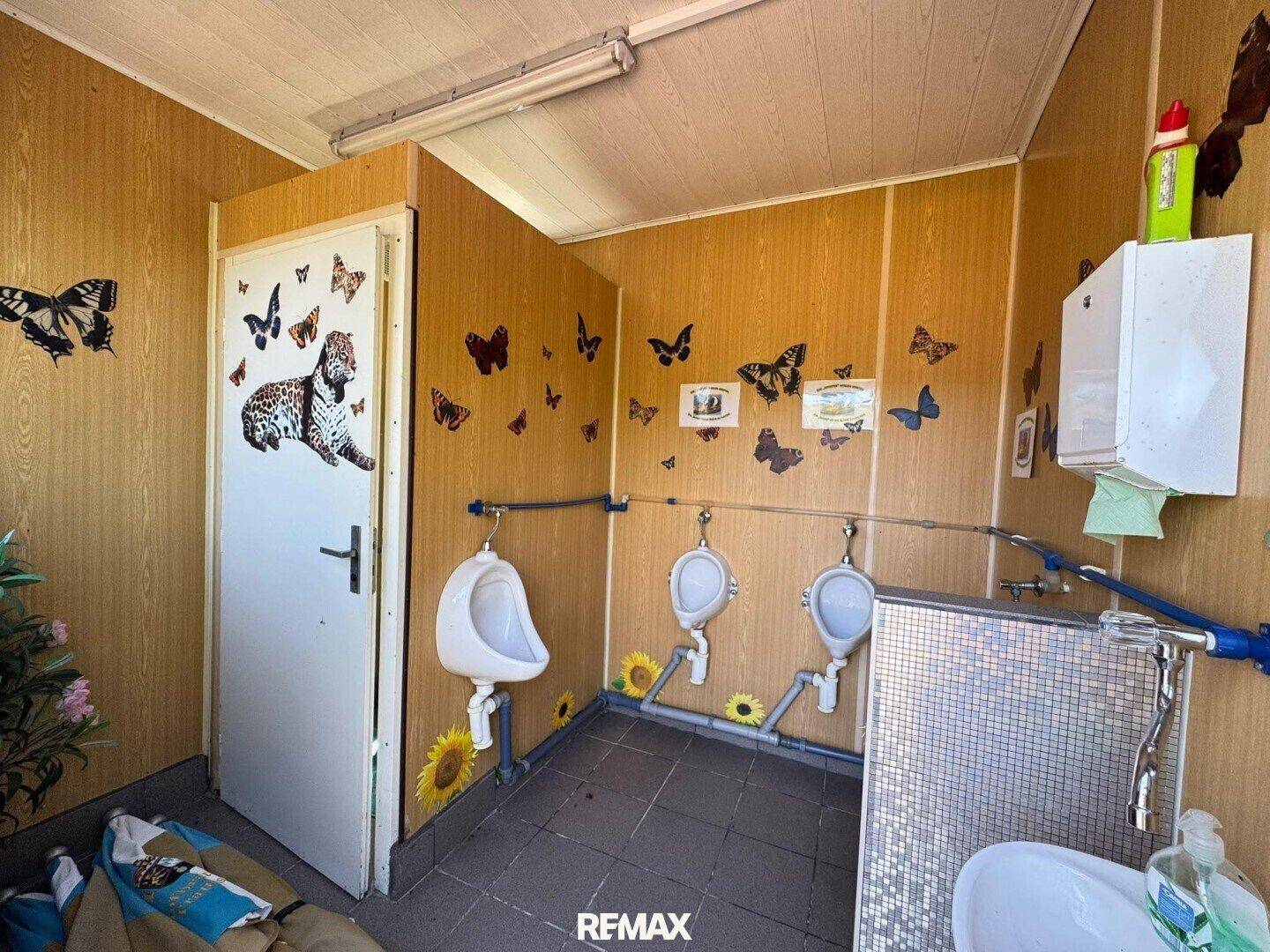 Herren-Toilette mit mehreren Urinalen und holzverkleideten Wänden, dekoriert mit Schmetterlingsmotiven.