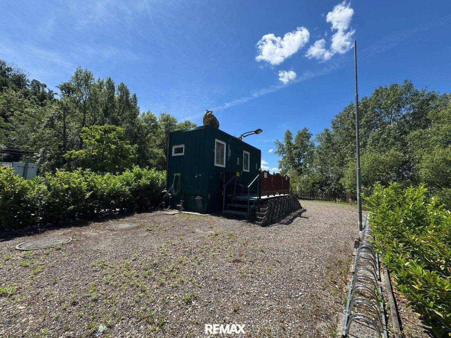 Grünes Containergebäude auf einem Schotterplatz, umgeben von Bäumen und Büschen unter blauem Himmel.