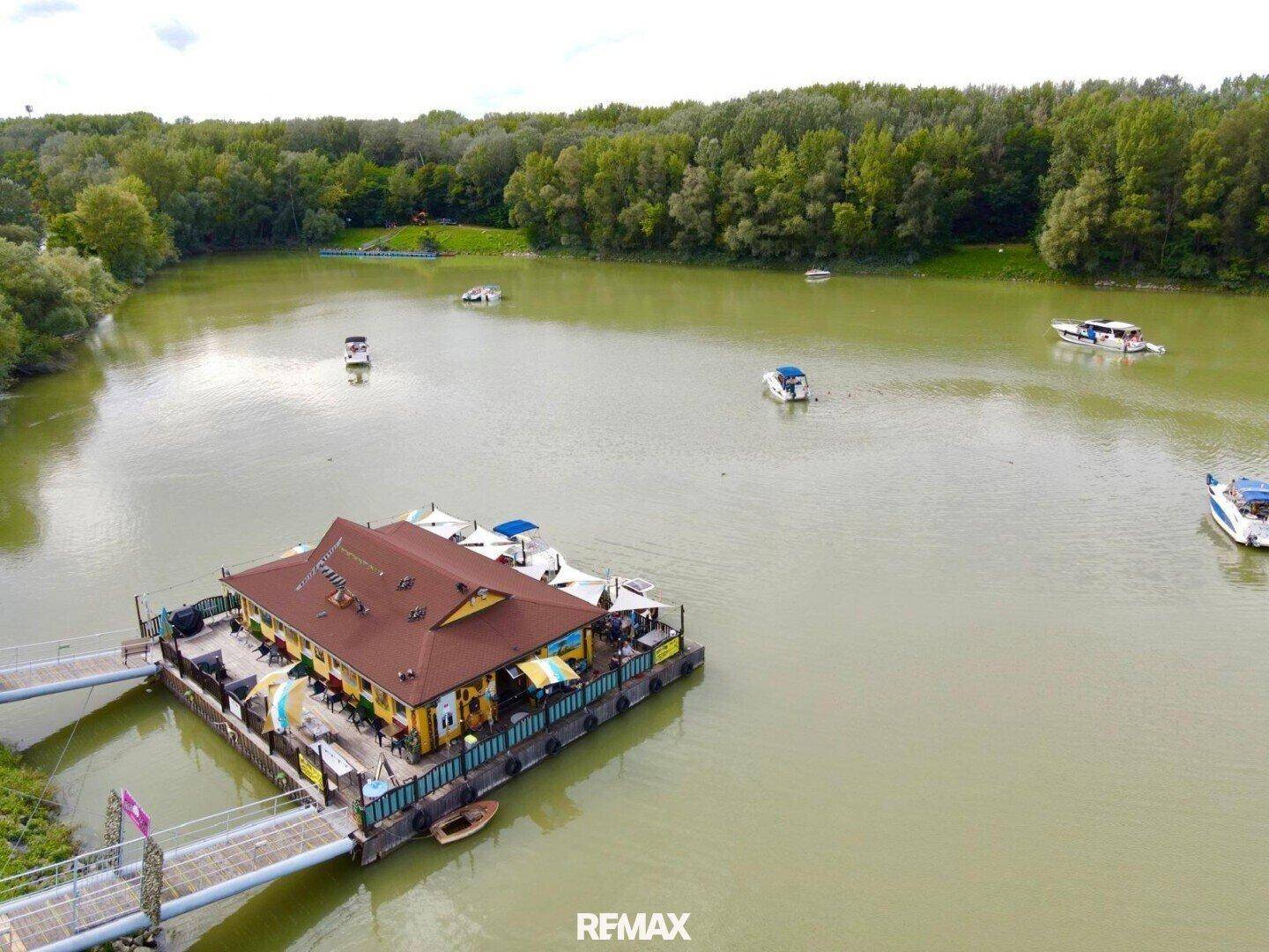 Eine weite Luftaufnahme des schwimmenden Restaurants auf dem See, umgeben von grünen Wäldern unter bewölktem Himmel.