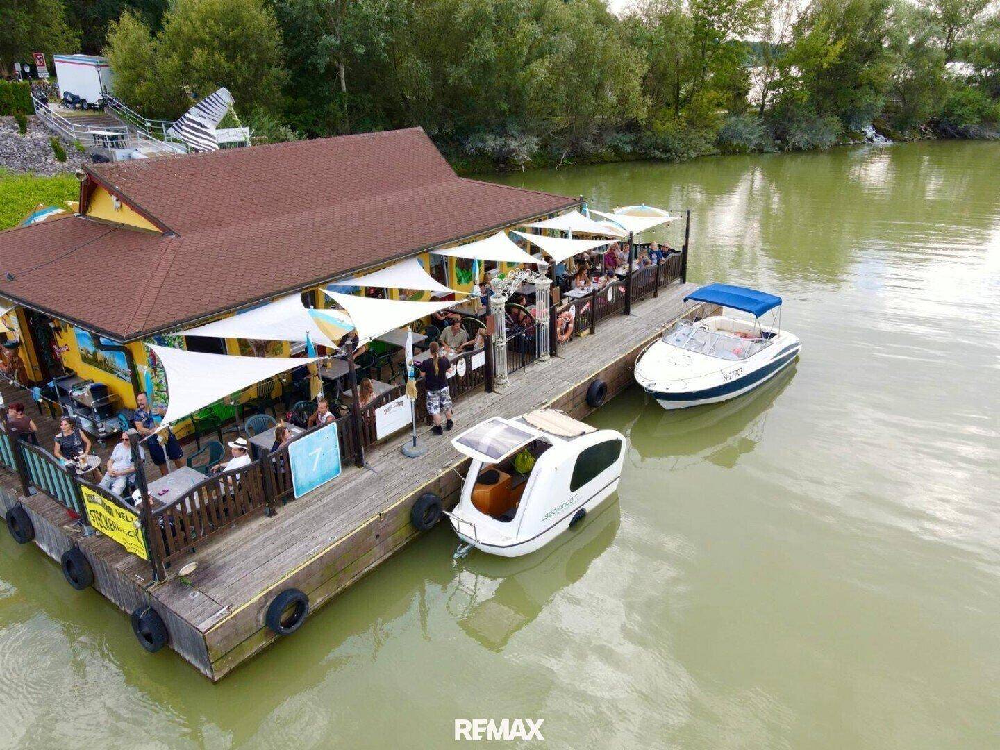 Nahaufnahme aus der Luft des schwimmenden Gebäudes mit Gästen auf der Terrasse und angedockten Sportbooten.