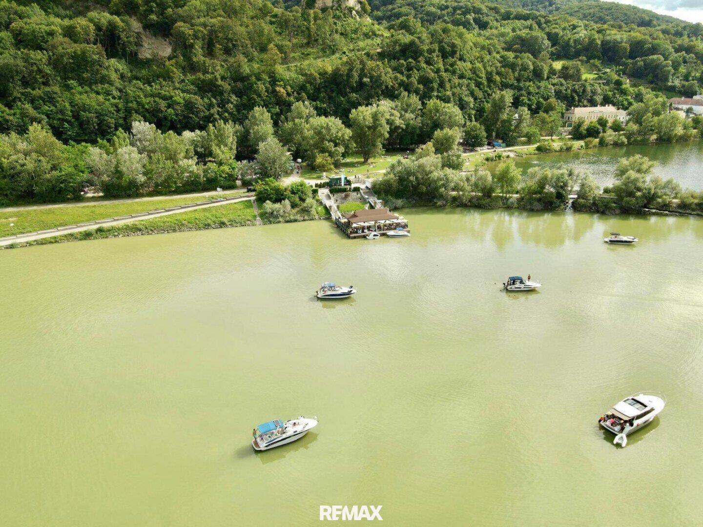 Luftaufnahme des Sees mit dem schwimmenden Gebäude, Booten und einem bewaldeten Hügel im Hintergrund.