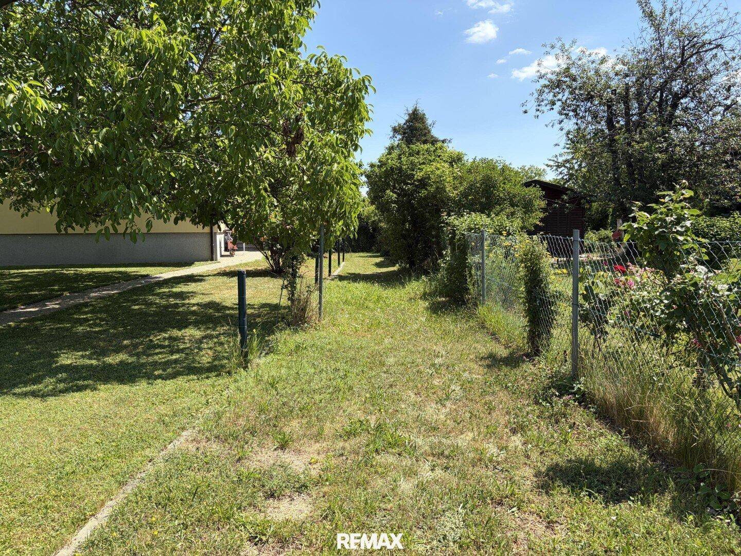 Ein langer Gartenweg mit einem Drahtzaun und verschiedenen Bäumen, der in die Ferne führt.