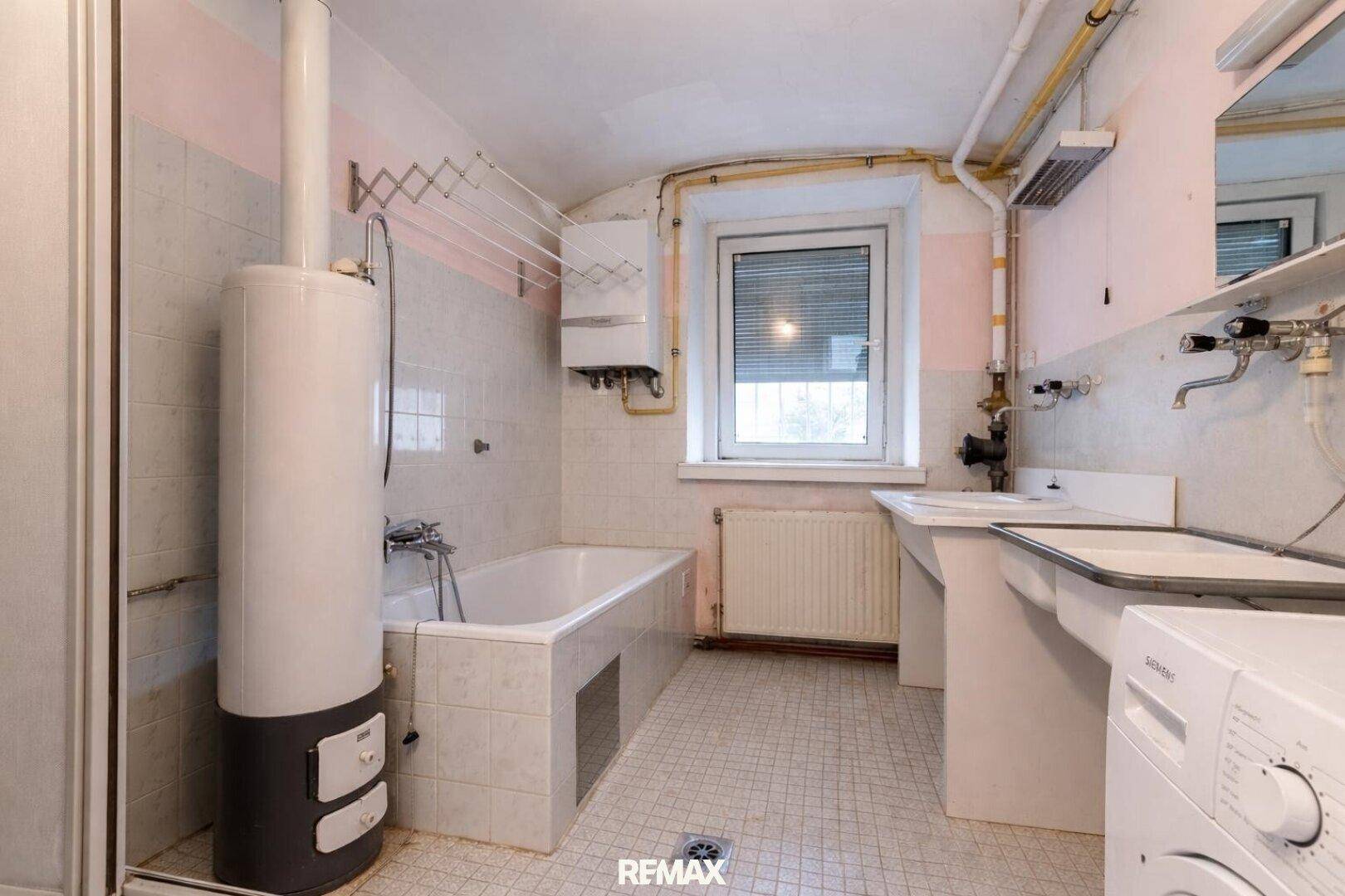 Älteres Badezimmer mit Badewanne, Waschbecken, Boiler und Fliesen in Pastellfarben, original erhalten.