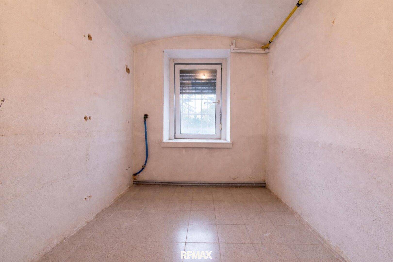 Leerer Raum mit Fliesenboden, einem Fenster und sichtbaren Wasserleitungen, in originalem Zustand.