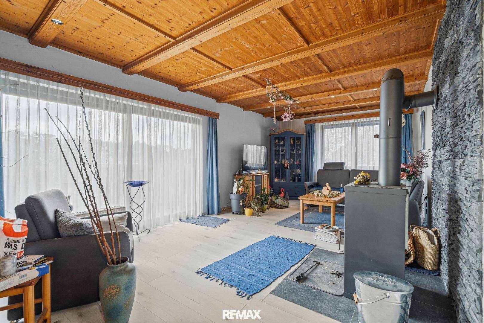 Helles Wohnzimmer mit Holzbalkendecke, Kaminofen, Steinwand und großen Fenstern.