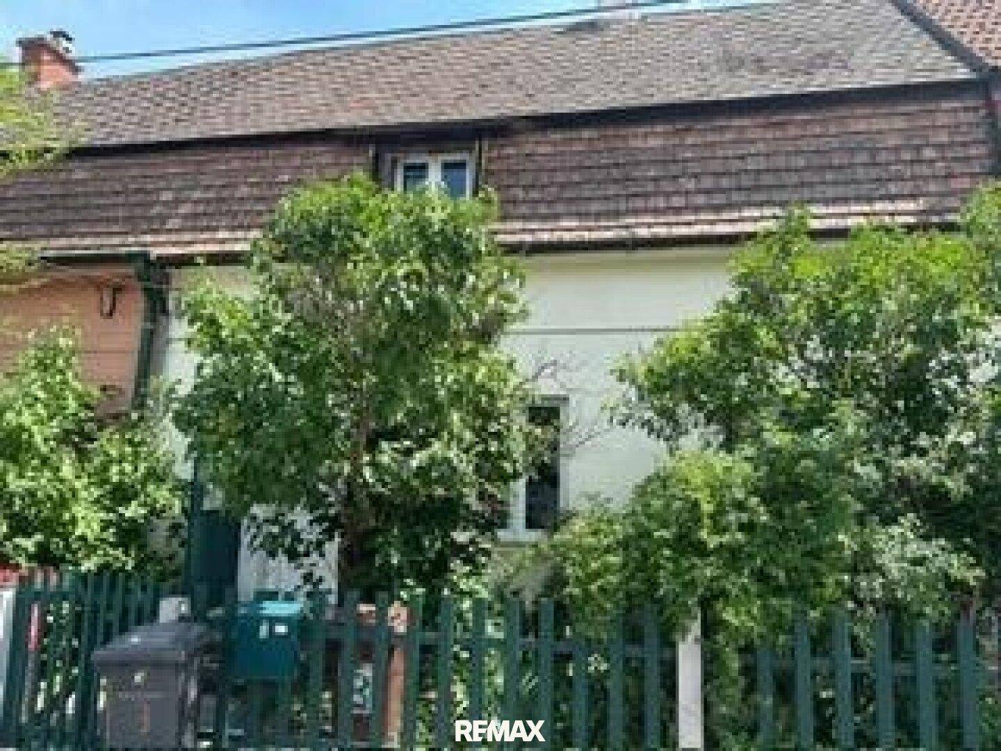 Vorderansicht eines älteren Hauses, teilweise von Bäumen und einem Zaun verdeckt.