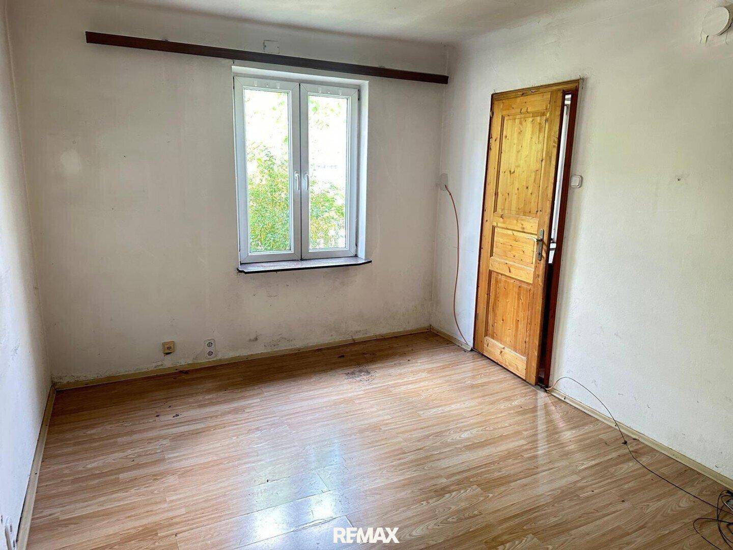 Leeres Zimmer mit Laminatboden, einem Fenster und einer Holztür, das Renovierung benötigt.