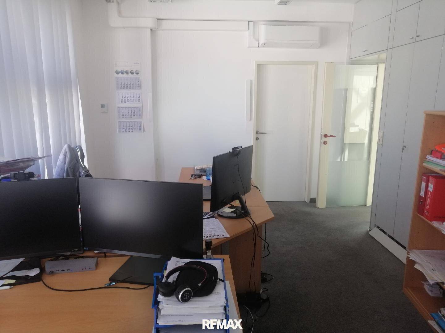 Geräumiges Büro mit zwei Arbeitsplätzen, Teppichboden und Klimaanlage an der Wand.