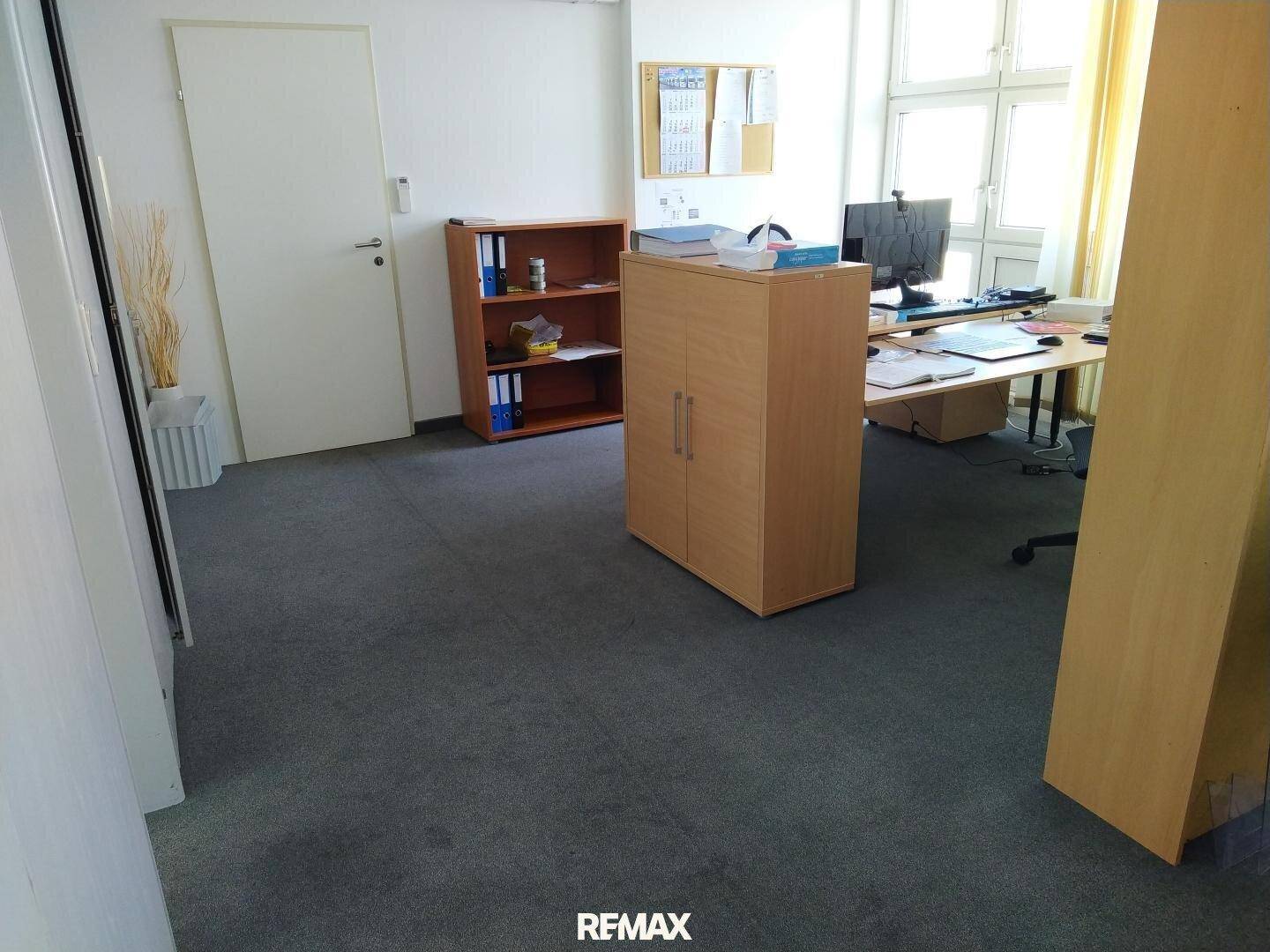 Helles Büro mit Teppichboden, Holzschränken und einem Arbeitsplatz am Fenster.