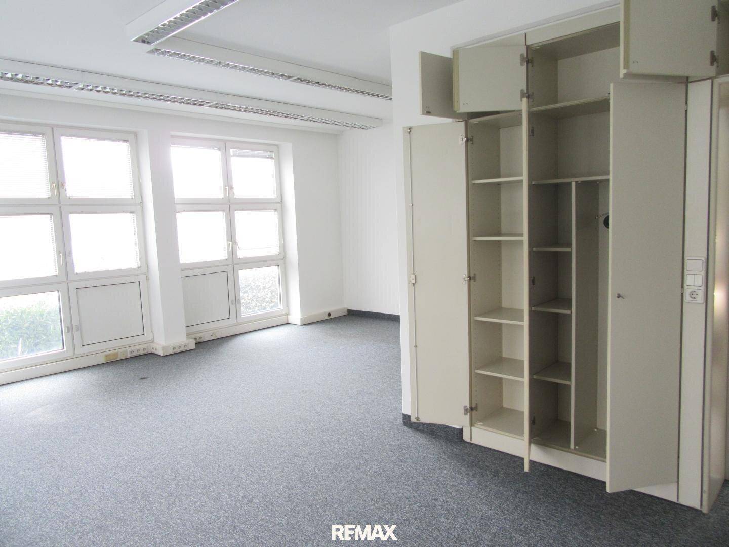 Geräumiger Büroraum mit Teppichboden, Einbauschränken und viel Tageslicht.