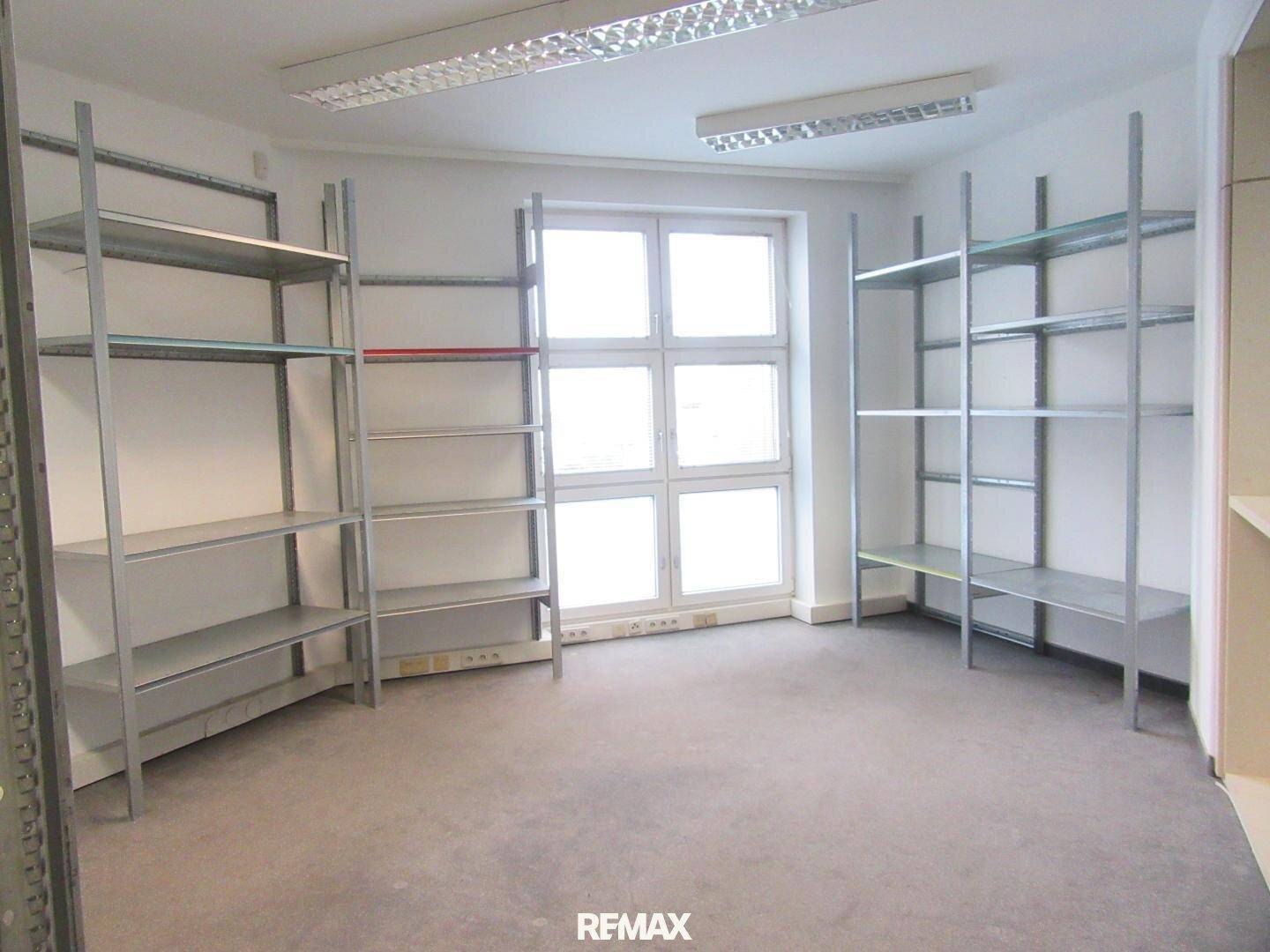 Funktionaler Büroraum mit Teppichboden, Metallregalen und großen Fenstern für Stauraum.
