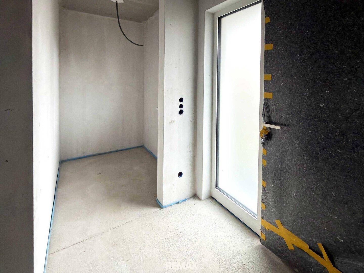 Kleiner Raum im Rohbau mit Estrichboden und einer Tür, ideal als Abstellraum oder Garderobe.