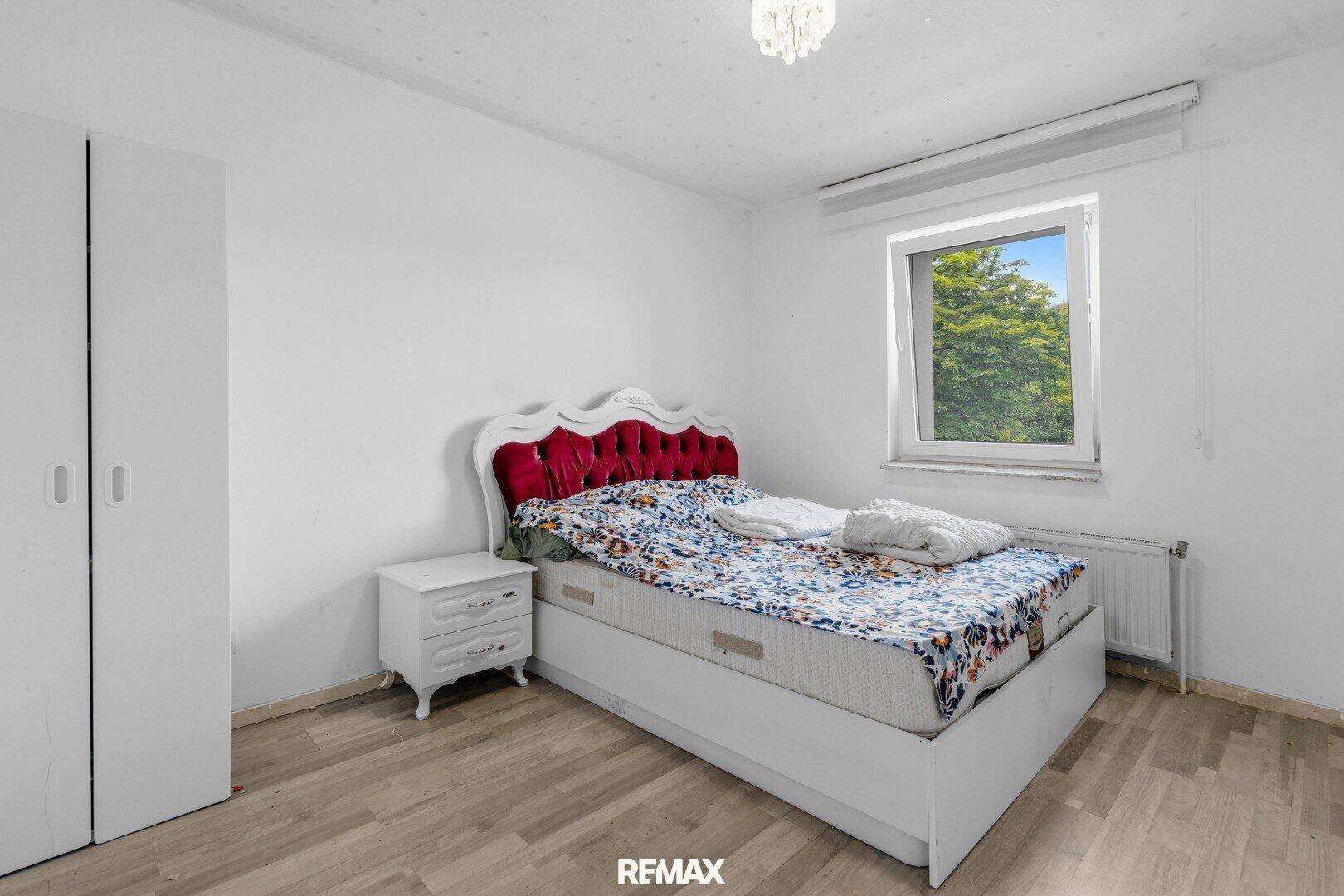 Schlafzimmer mit rotem Bett, weißem Nachttisch und Fenster mit Blick ins Grüne.