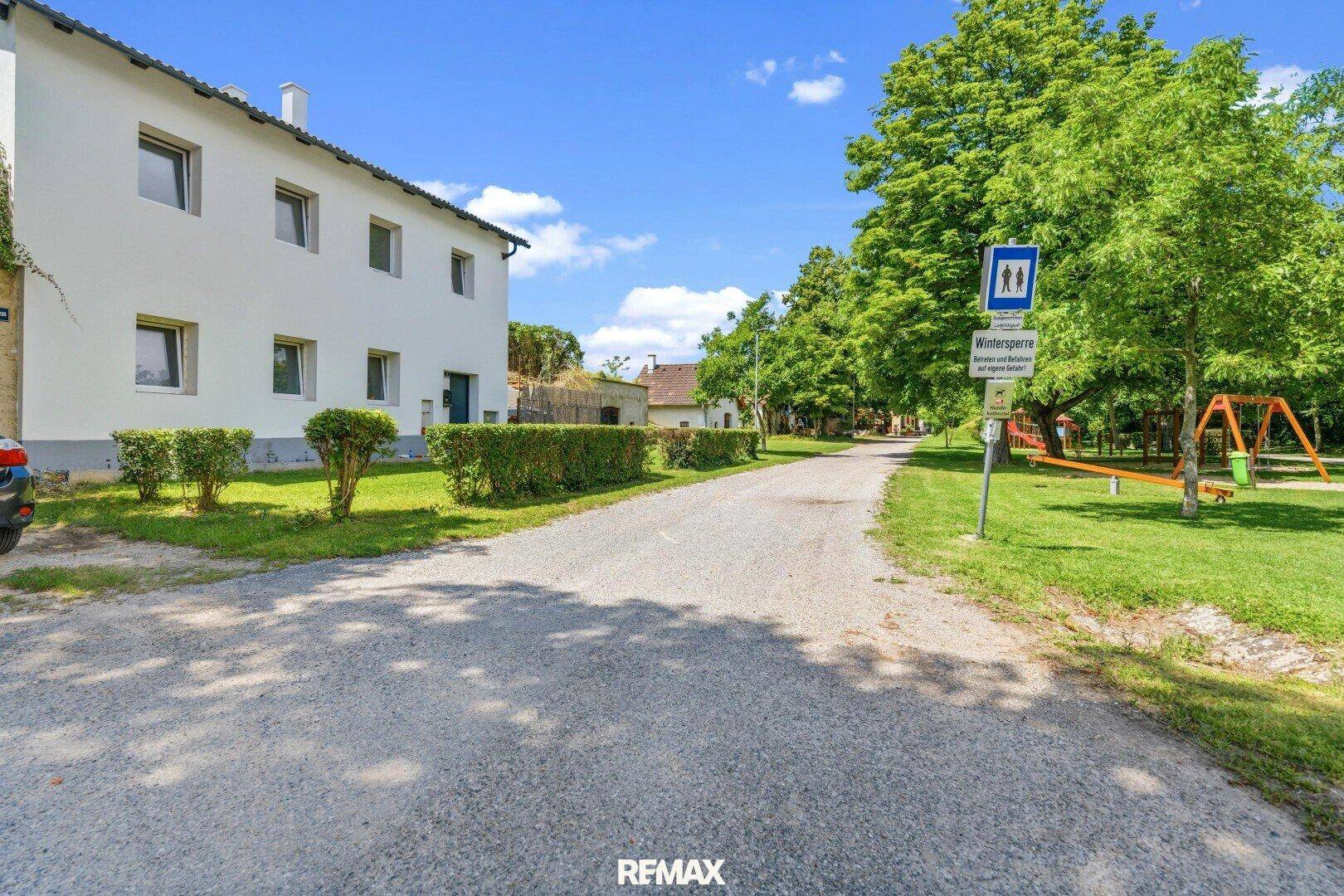 Kiesweg führt zu einem Spielplatz neben dem Haus mit viel Grün und Bäumen unter blauem Himmel.