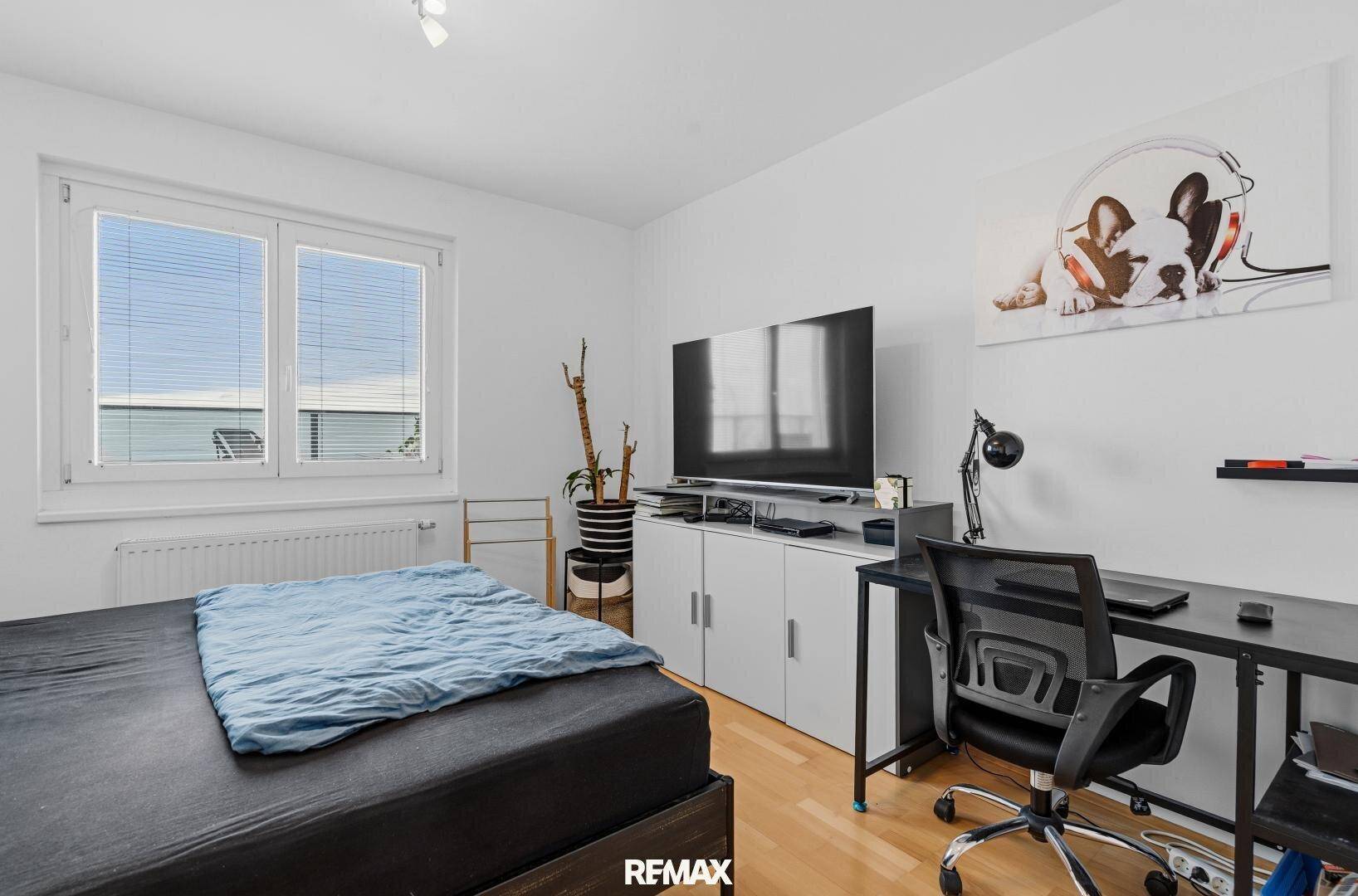Schlafzimmer mit Bett, Fernseher und Schreibtisch, mit hellem Holzboden.