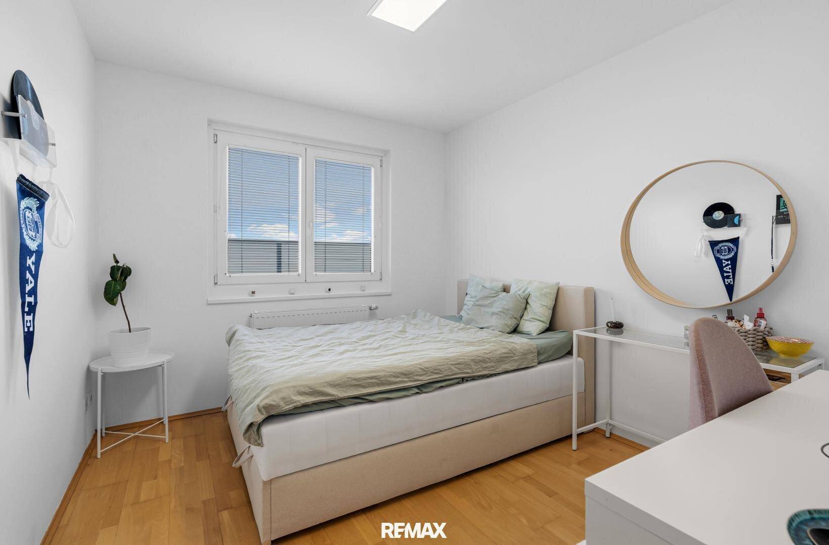 Helles Schlafzimmer mit Bett, Schreibtisch und einem runden Spiegel, schlicht eingerichtet.