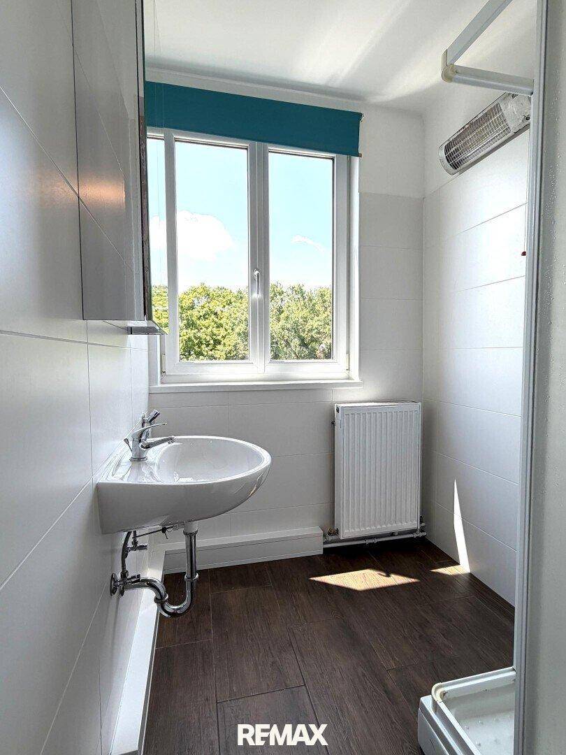 Helles Badezimmer mit Dusche, Waschbecken und Fenster mit Blick ins Grüne.