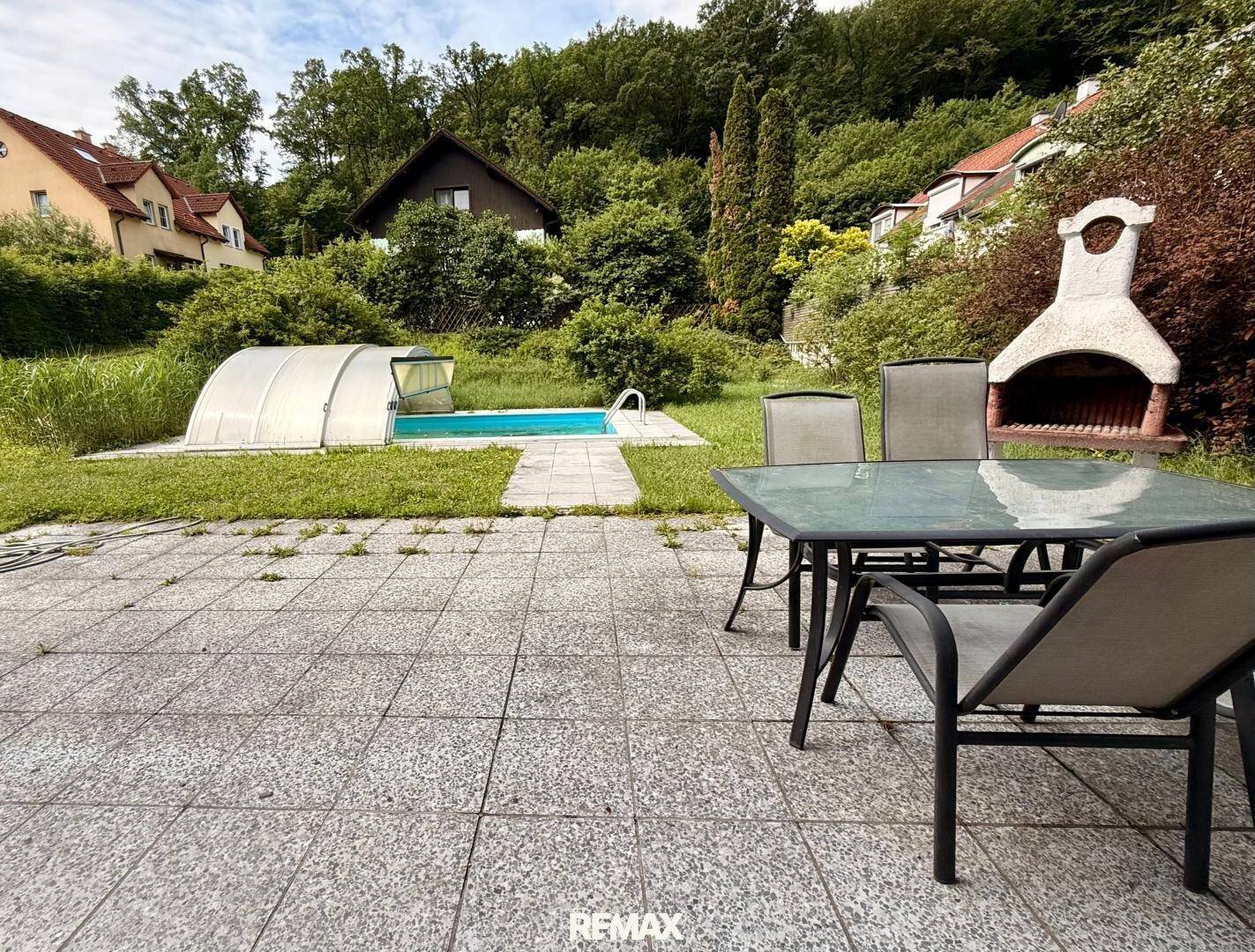 Große Steinterrasse mit Gartenmöbeln, Grillplatz und Blick auf den Poolbereich und die grüne Umgebung.