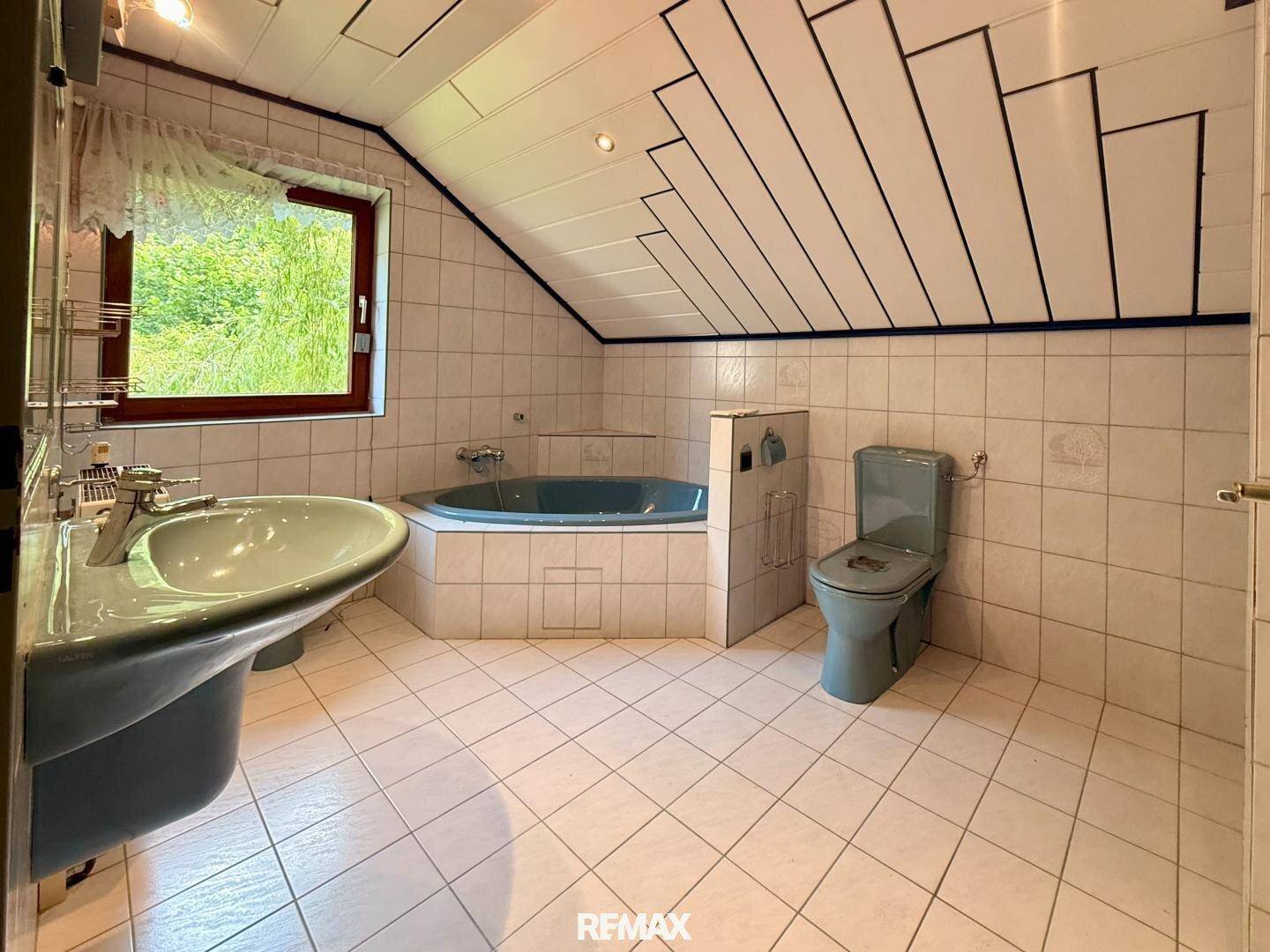 Badezimmer im Dachgeschoss mit Eckbadewanne, Fliesenboden und Fenster, im Stil der 80er Jahre.
